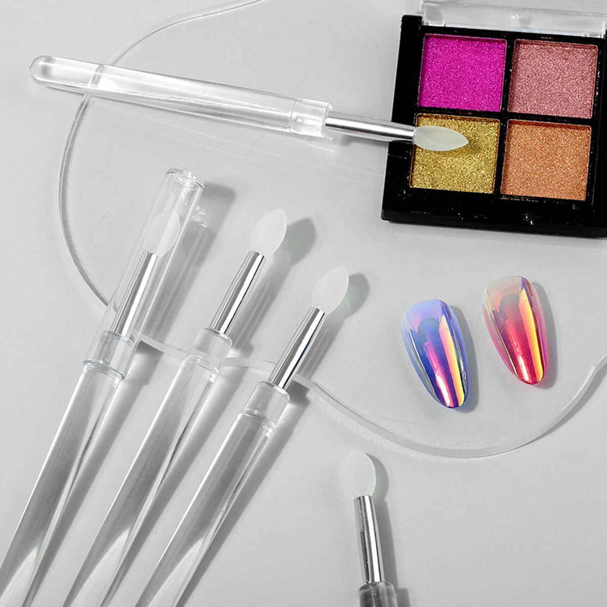 Plusieurs applicateurs transparents à embouts en silicone à côté d'un quad de fard à paupières et d'ongles holographiques.