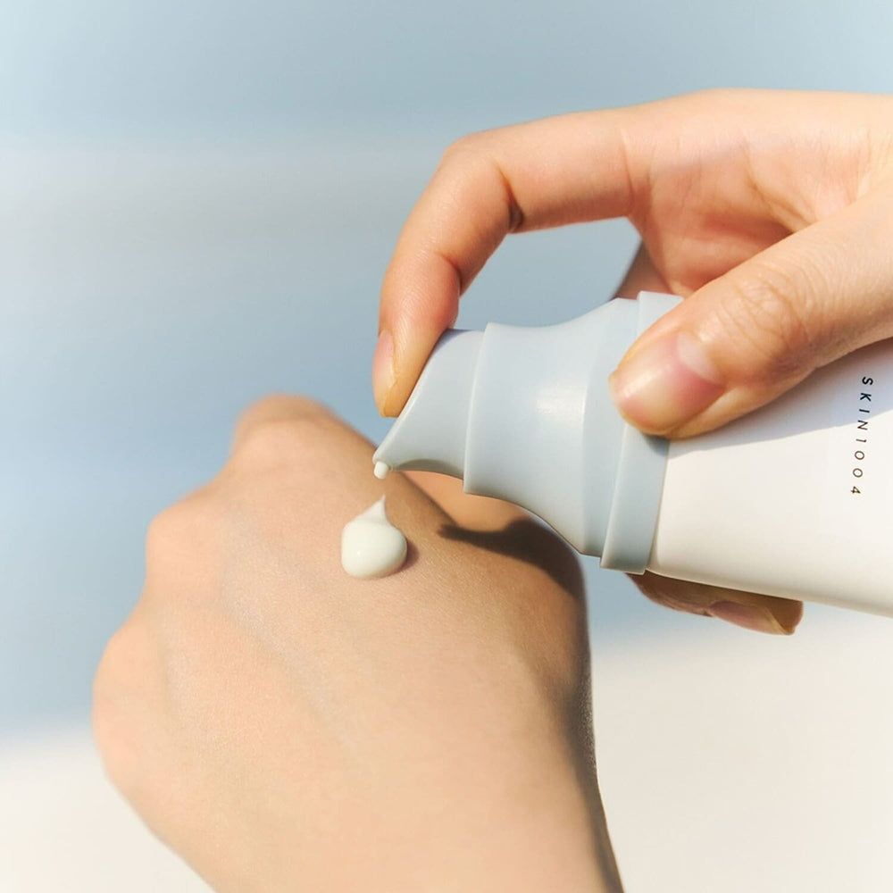 Main pressant de la crème d'un tube. Texte: SKIN1004. La crème est blanche. Le fond est bleu.