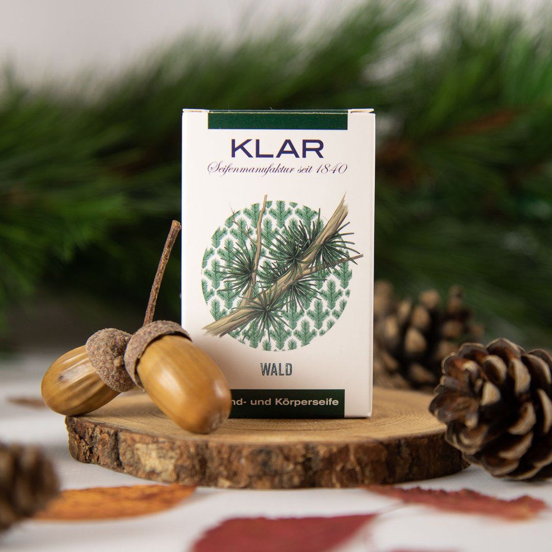 Klar zeepverpakking op houten schijf met eikels en dennenappels. Verpakking: wit met groen logo en takillustratie.