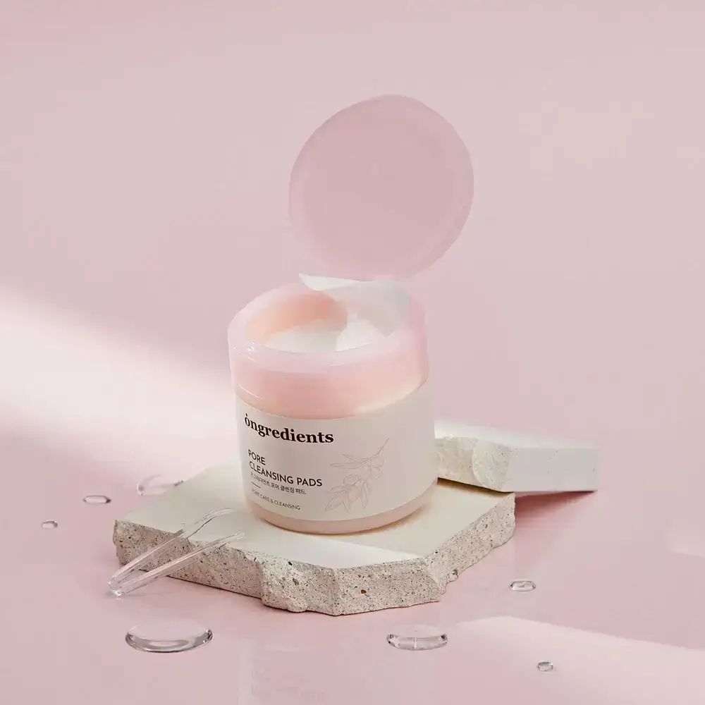Ronde pot met roze open deksel. Opschrift: Ongredients, Pore Cleansing Pads. Geplaatst op steen.