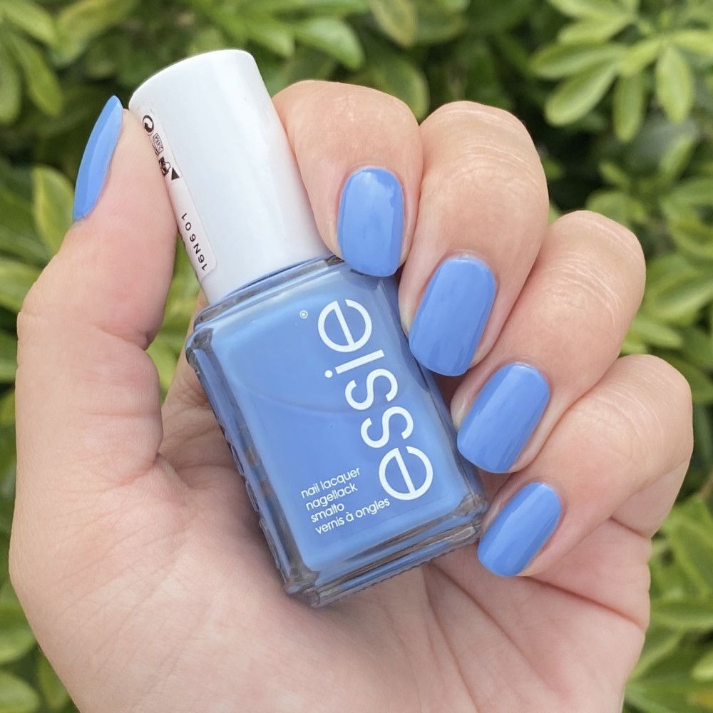 Essie nagellakflesje en gelakte nagels in lichtblauw. Flesje met witte dop, etiket met tekst. Hand houdt flesje vast.