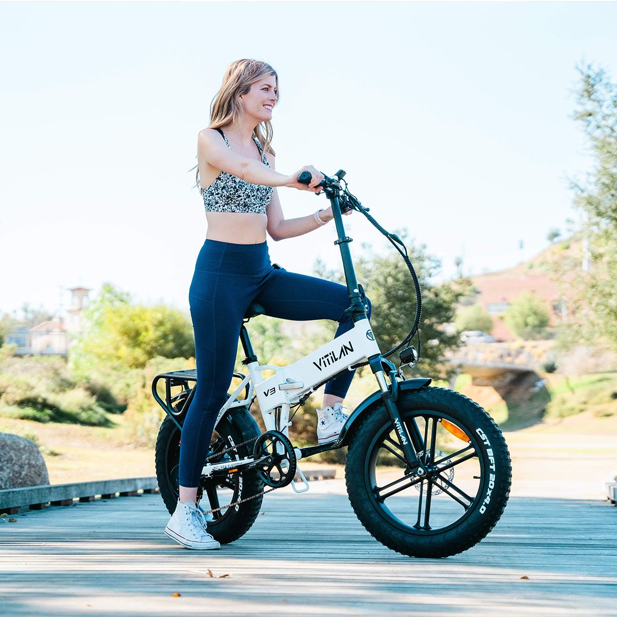 Vélo électrique pliable blanc avec pneus noirs. Une femme fait du vélo sur un pont. Vitilan V3 est écrit sur le cadre.