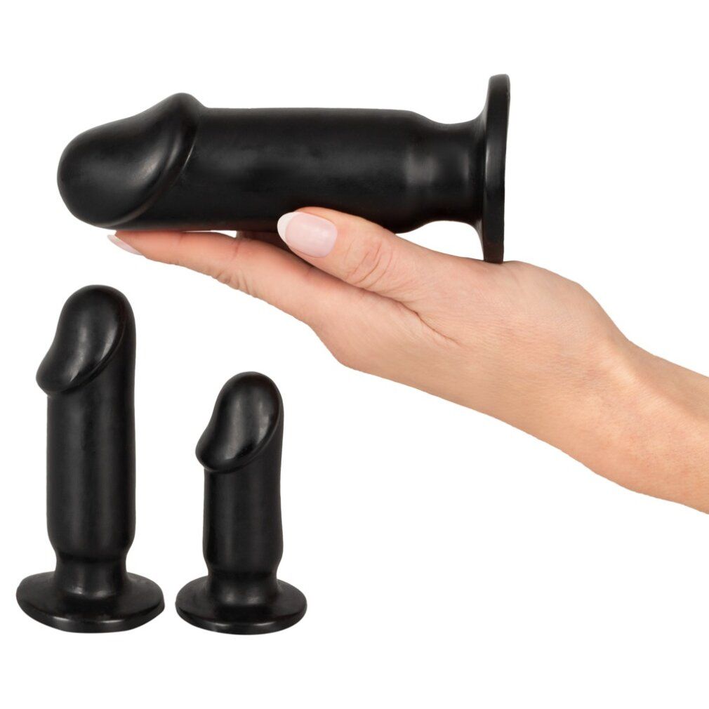 Trois plugs anaux noirs. Un plug est tenu par une main. Les deux autres sont à côté. Forme cylindrique avec pointe arrondie.
