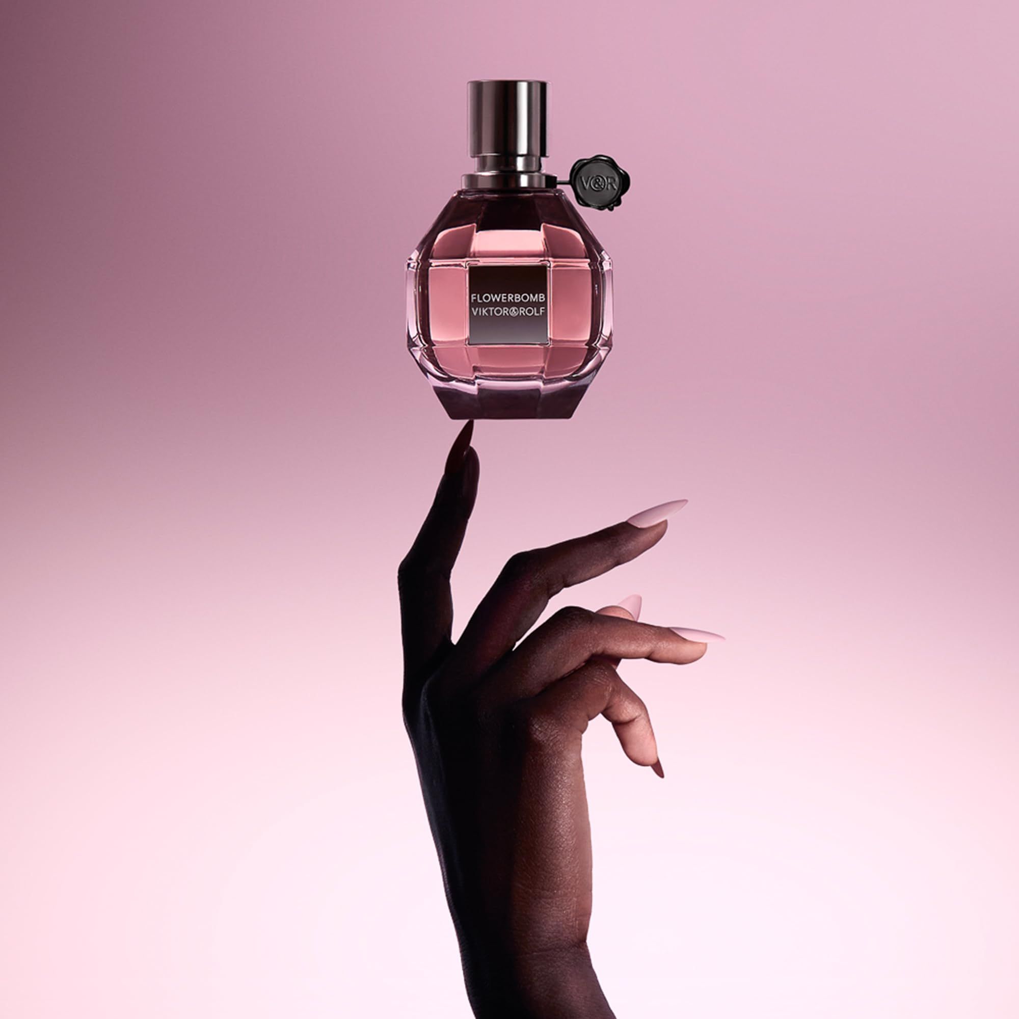 Flacon de parfum Flowerbomb de Viktor & Rolf tenu par une main avec de longs ongles clairs. Fond rose.