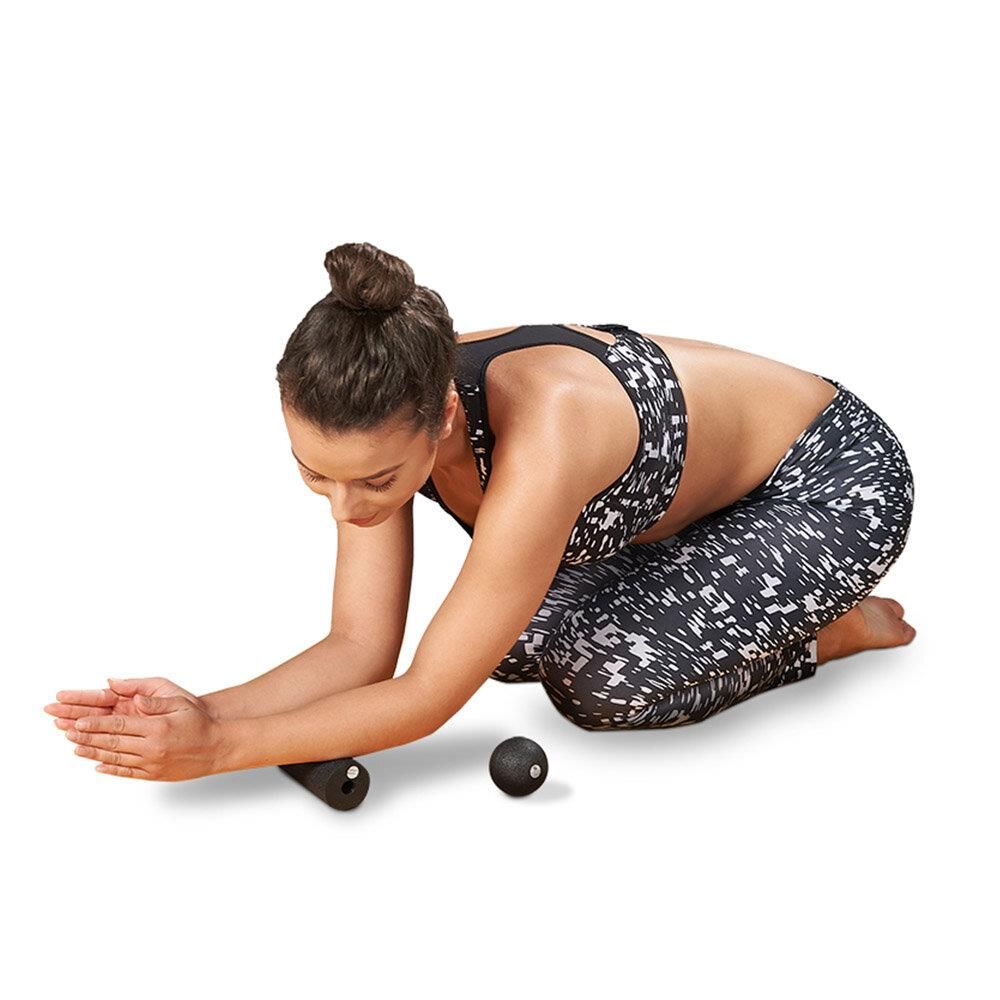 Vrouw in sportkleding knielt met fascia roller en bal. Ze steunt op haar handen, blik naar beneden.
