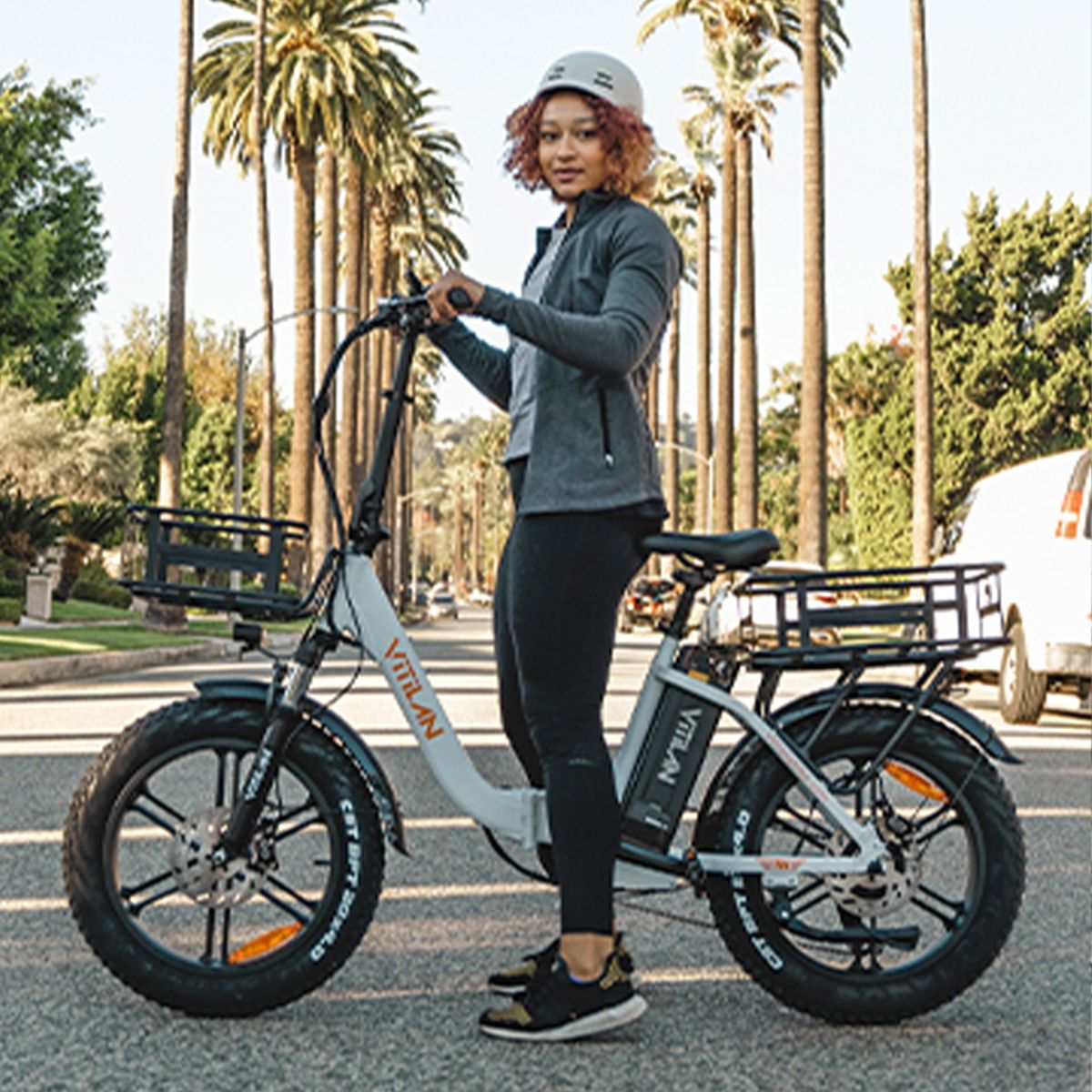Personne debout à côté d'un vélo électrique Vitilan blanc. Elle porte un casque et des vêtements de sport. Palmiers en arrière-plan.