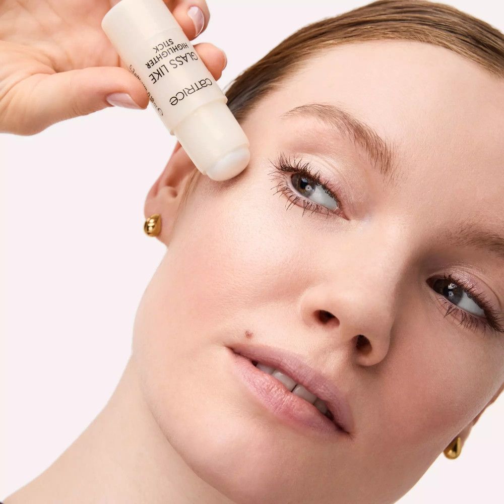 Hand houdt highlighter stick op het gezicht van een vrouw. Opschrift: Catrice, GLASS LIKE HIGHLIGHTER STICK.