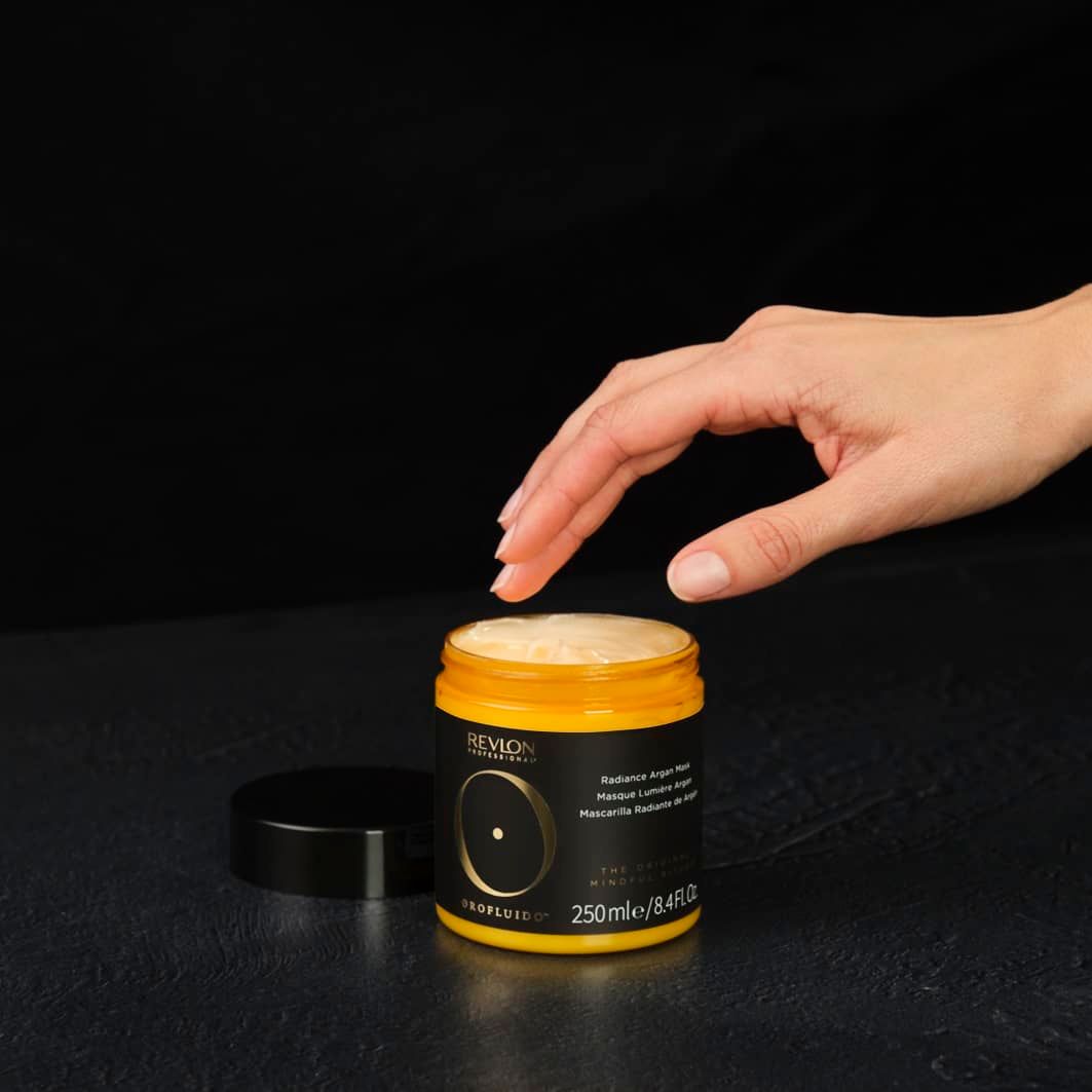 Main touchant le pot ouvert. Étiquette jaune. Inscription: Revlon, Orofluido, Radiance Argan Mask. 250 ml.