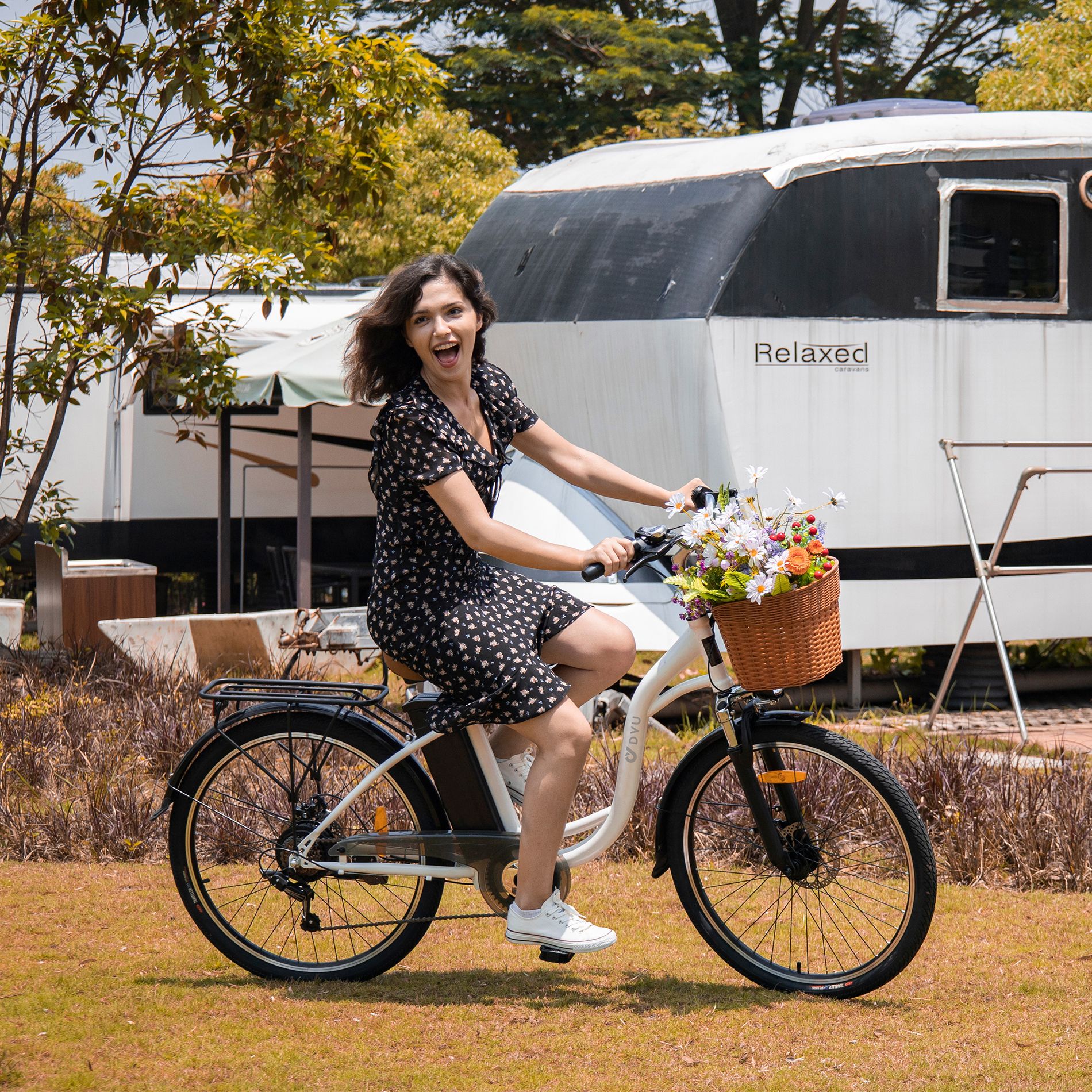 Vrouw op witte fiets met mand en bloemen. Zwart frame. Achtergrond: gebouw.