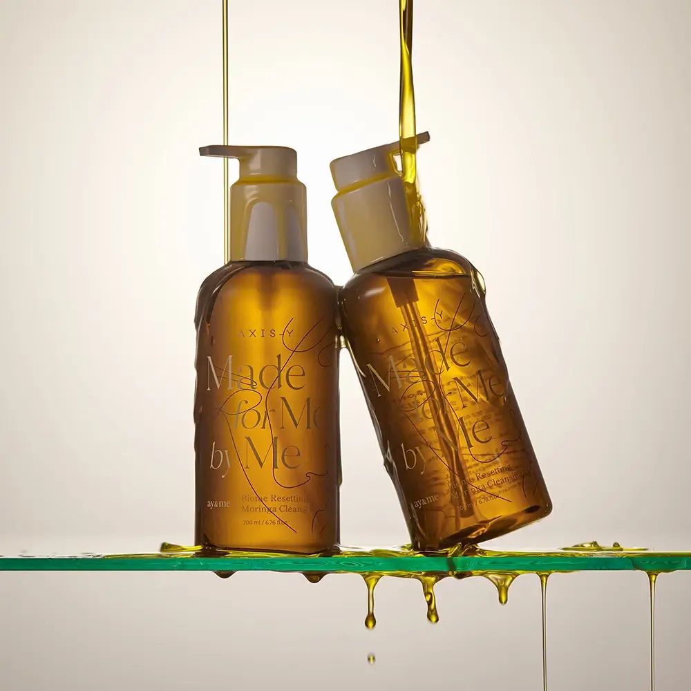 Twee bruine flessen met witte pompen. Olie druppelt over de flessen. Opschrift: AXIS-Y, Made for Me by Me.