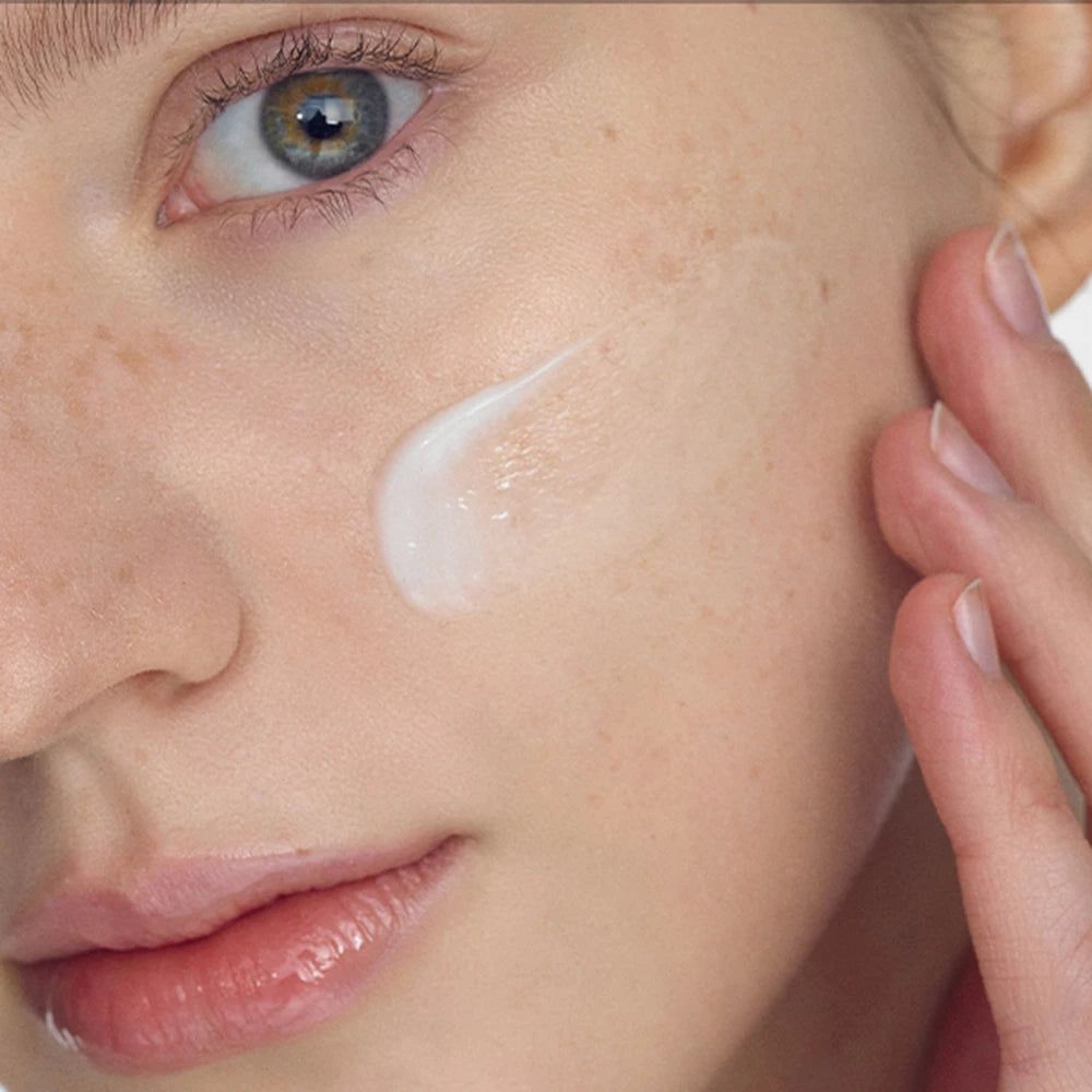 Close-up van een gezicht met crème op de wang. Een hand raakt de huid aan. De crème is wit.