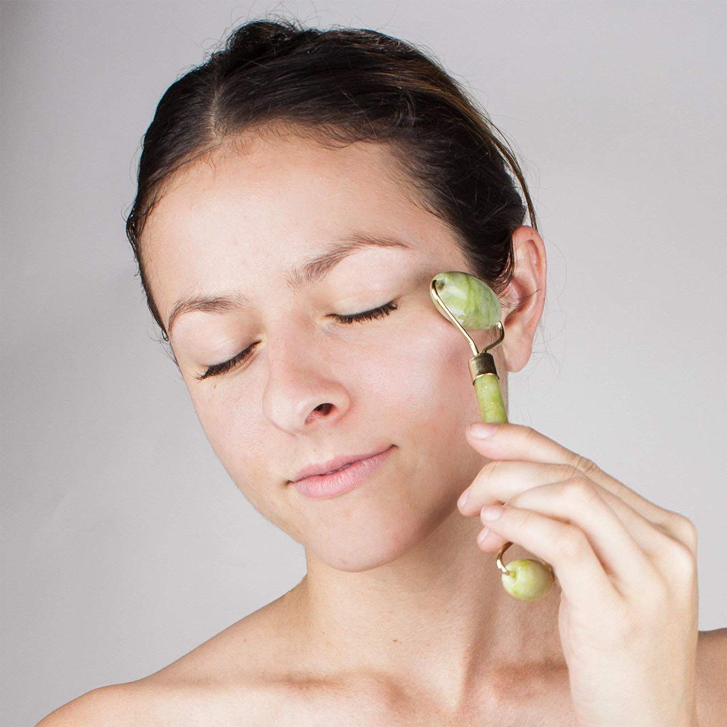 Une femme aux yeux fermés utilise un rouleau facial en jade vert sur sa tempe.
