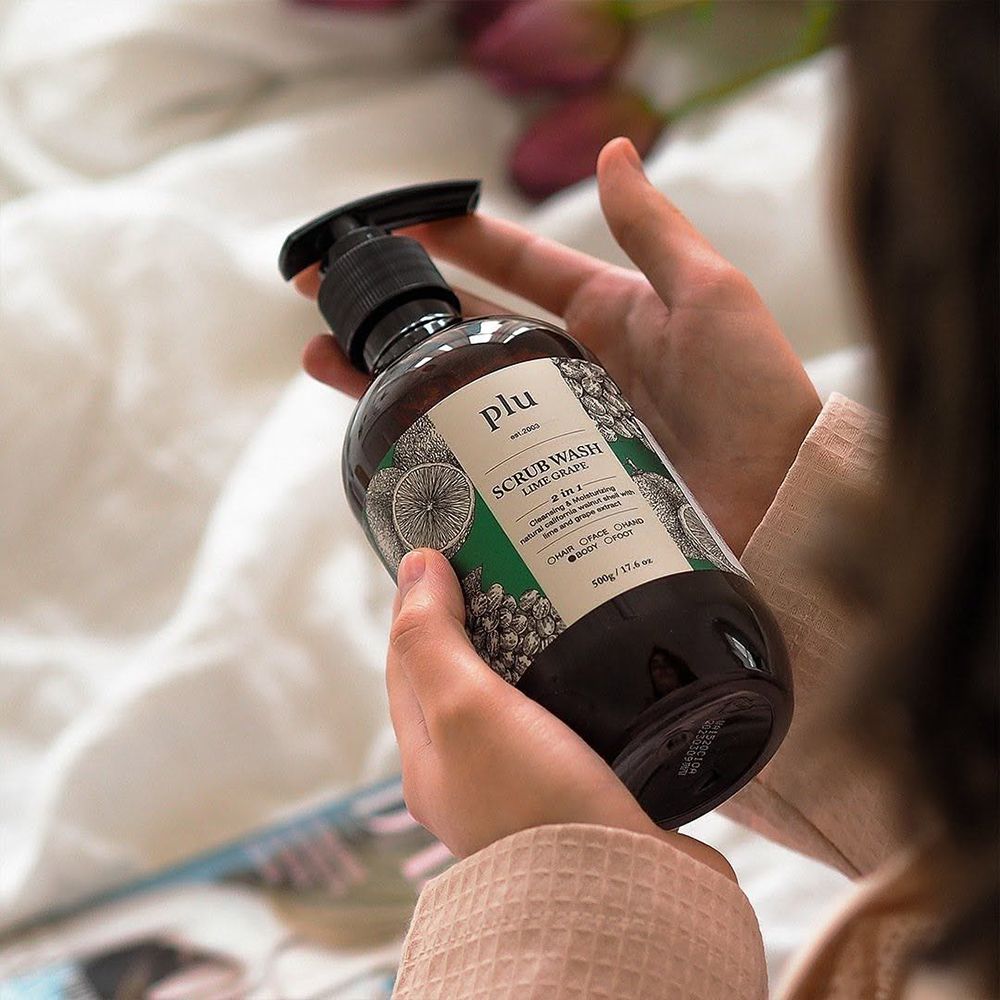 Flacon brun avec pompe noire. Inscription : PLU Scrub Wash Lime Grape. Mains tenant le flacon.