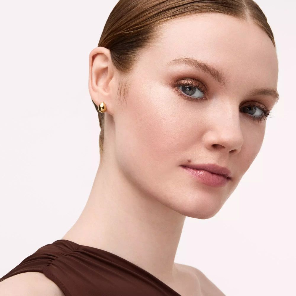 Portrait d'une femme avec fard à paupières marron. Elle porte des boucles d'oreilles dorées. Fond clair.