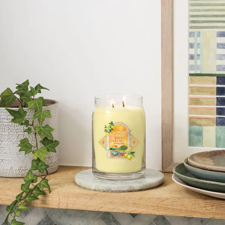 Grand pot de bougie jaune avec deux mèches et étiquette. Sur dessous de plat en marbre. Fond : étagère, plantes, décoration.