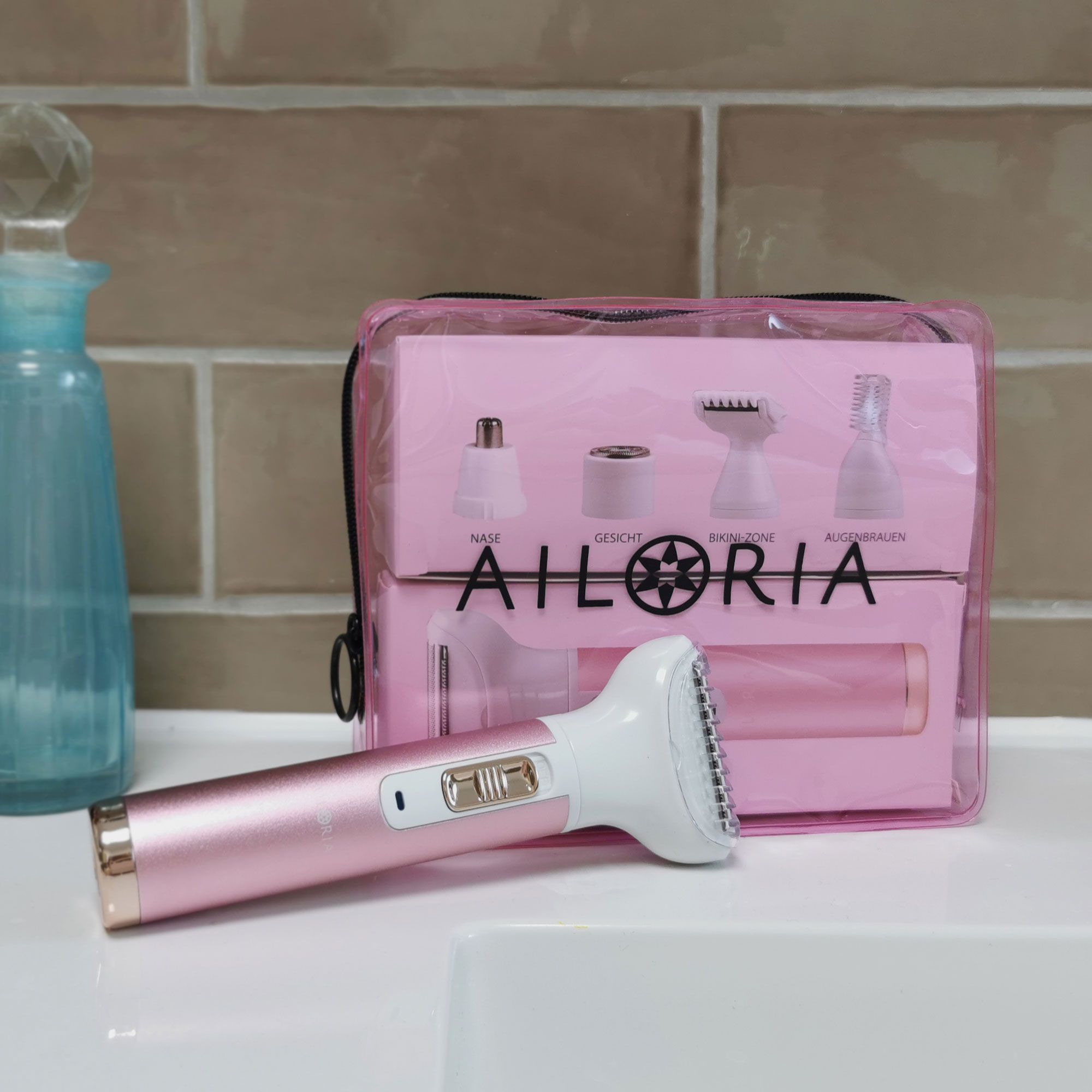 Rasoir et trousse sur un lavabo. Logo AILORIA. Têtes représentées. Inscriptions: VISAGE, ZONE BIKINI, SOURCILS. Trousse rose.
