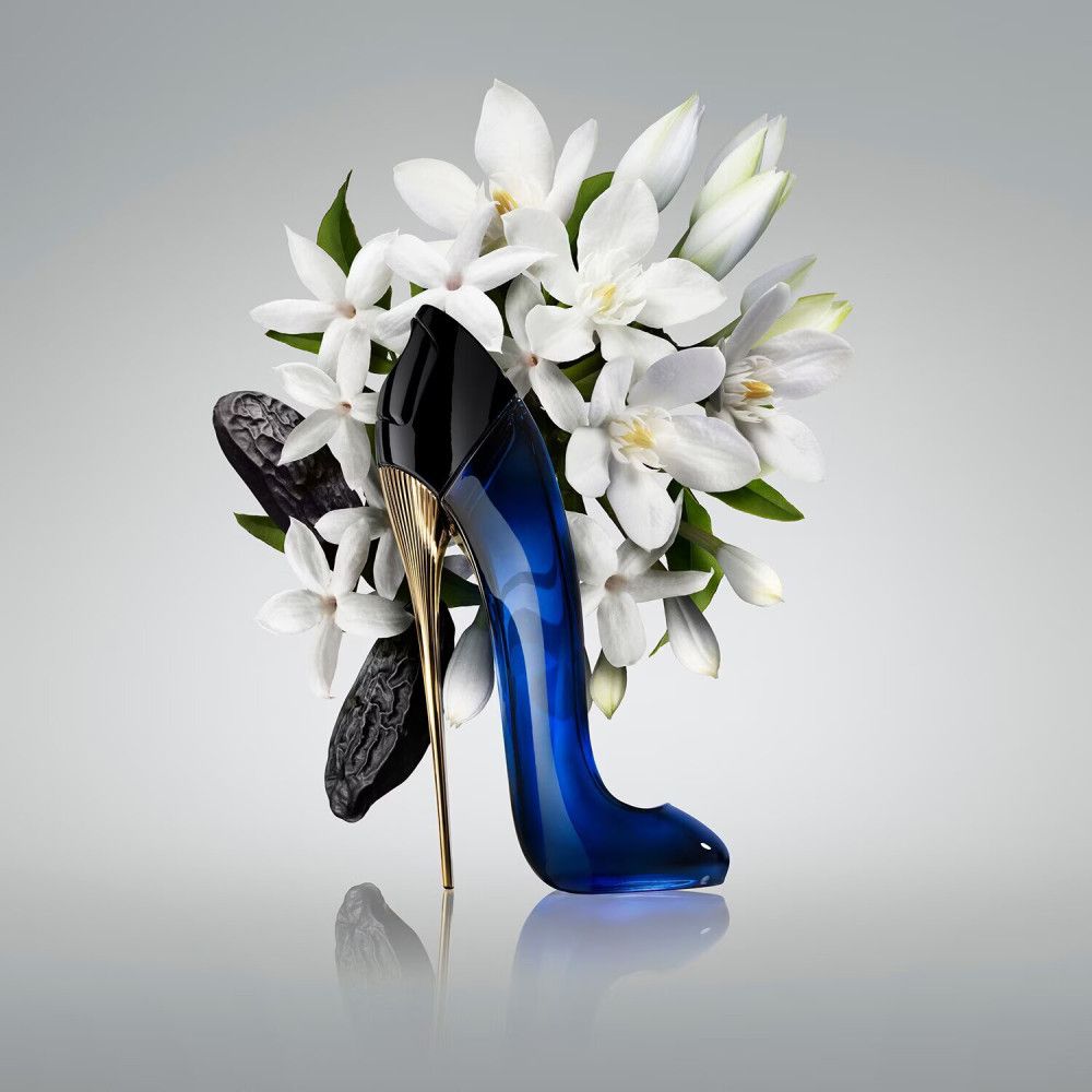 Flacon bleu en forme d'escarpin à talon doré, entouré de fleurs blanches et de fèves sombres.