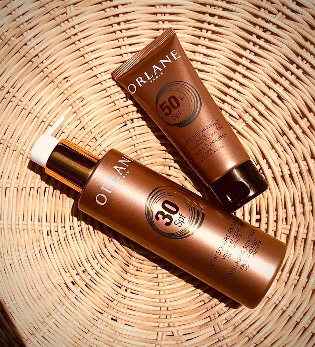 Deux crèmes solaires. Une bouteille 30 SPF et un tube 50 SPF. Bruns avec détails dorés. Sur un fond en osier.