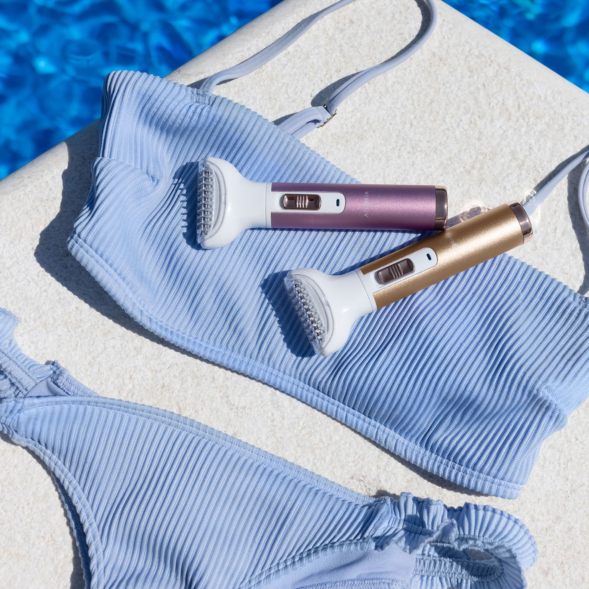 Twee scheerapparaten met witte kop en gekleurde handvatten. Liggend naast een blauwe bikini aan de zwembadrand.