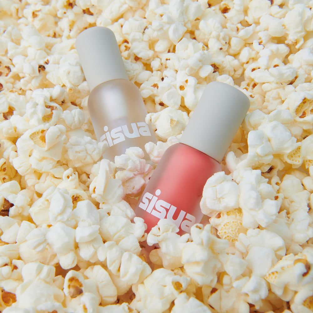 Twee flessen lipgloss, omringd door popcorn. De flessen zijn transparant met witte doppen. Gedrukte tekst: sisuia.