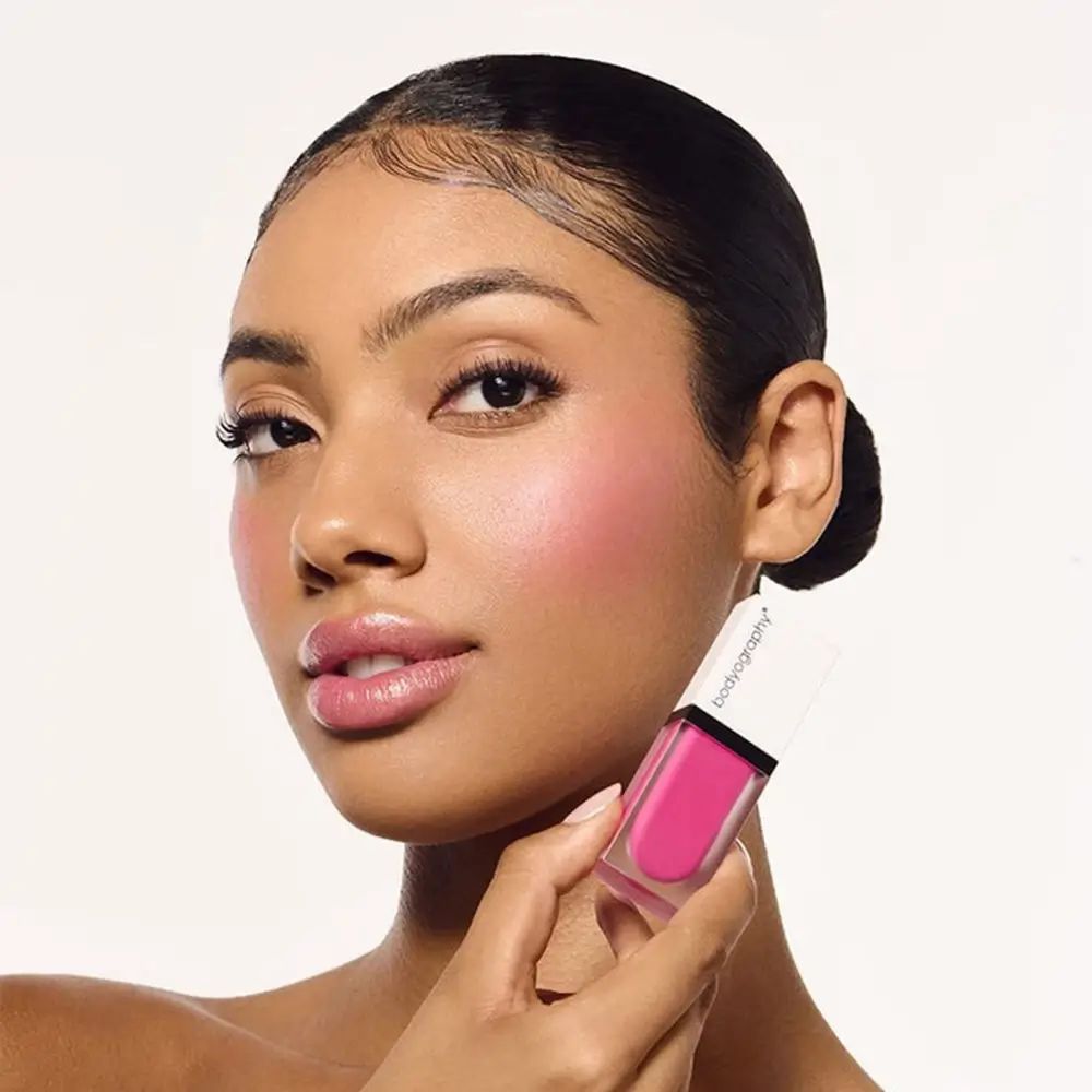 Vrouw met roze blush op de wangen. Houdt een flesje Bodyography vloeibare blush vast.