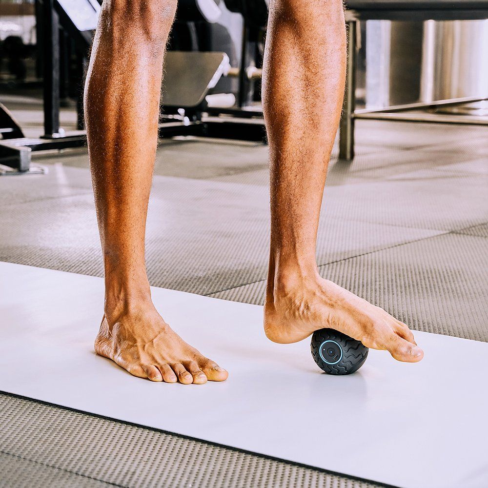 Pieds sur un tapis, une balle de massage noire sous le pied. Environnement de salle de sport.