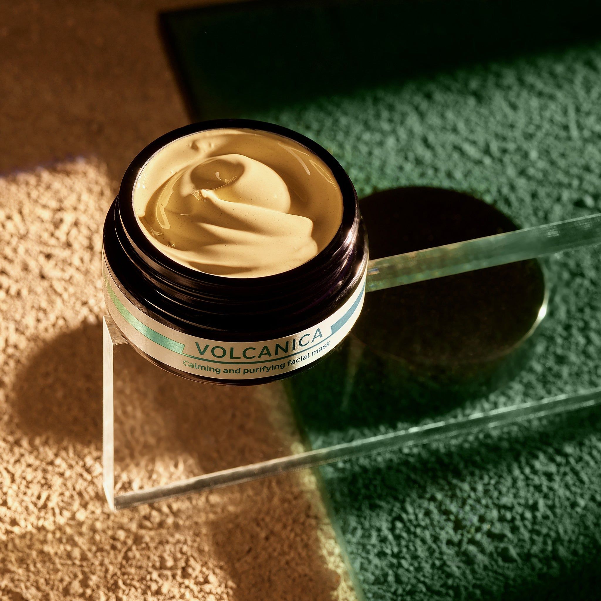 Open pot met romig masker. Opschrift: VOLCANICA, Calming and purifying facial mask. Op glazen plaat.