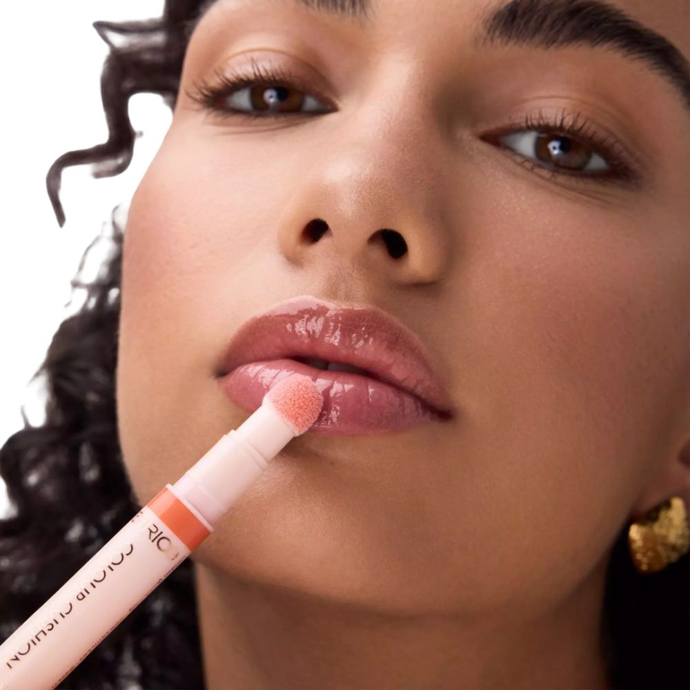 Vrouw brengt Catrice Colour Cushion Juicy Lippenöl aan. Roze tube met applicator. Tekst: Colour Cushion Juicy Lip Oil.