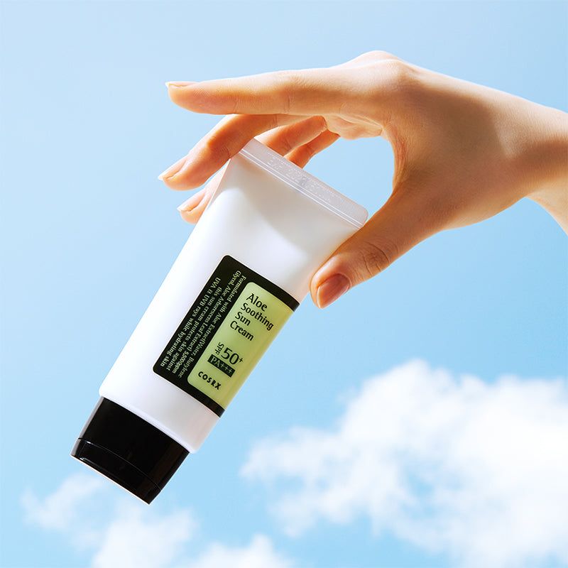 Hand houdt tube vast. Tekst: Aloe Soothing Sun Cream SPF50+ PA+++. Zwarte dop. Blauwe hemel.