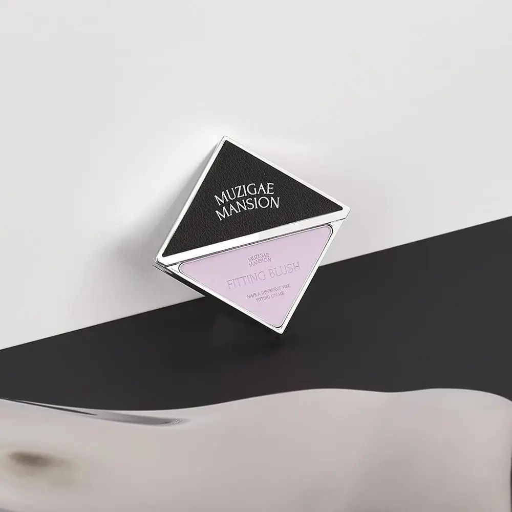 Vierkante rougeverpakking op zwart-witte achtergrond. Zwart deksel met MUZIGAE MANSION. Roze rouge in driehoekige vorm.