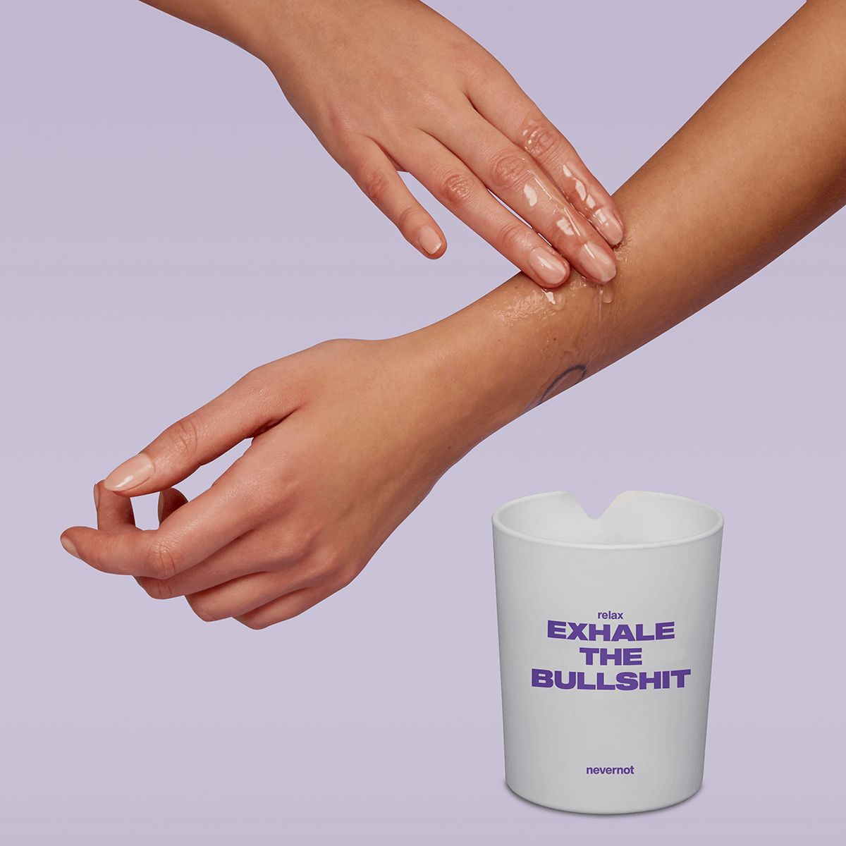 Handen wrijven vloeistof op arm. Kaarsbeker met opschrift "relax EXHALE THE BULLSHIT". Merk: nevernot. Achtergrond: lichtpaars.