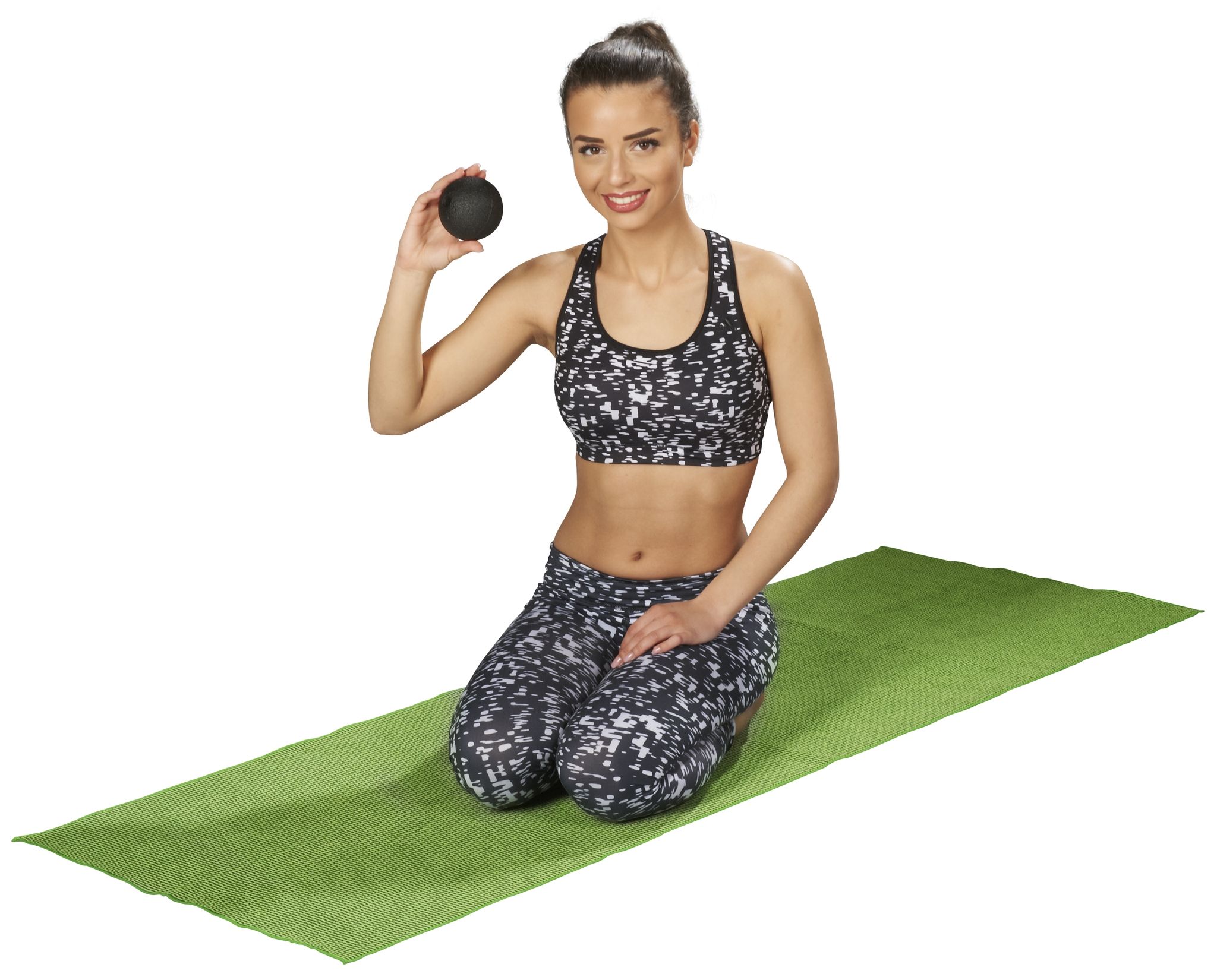 Femme assise sur un tapis vert. Tient une balle noire. Vêtements de sport : haut et pantalon à motifs. Sourit.