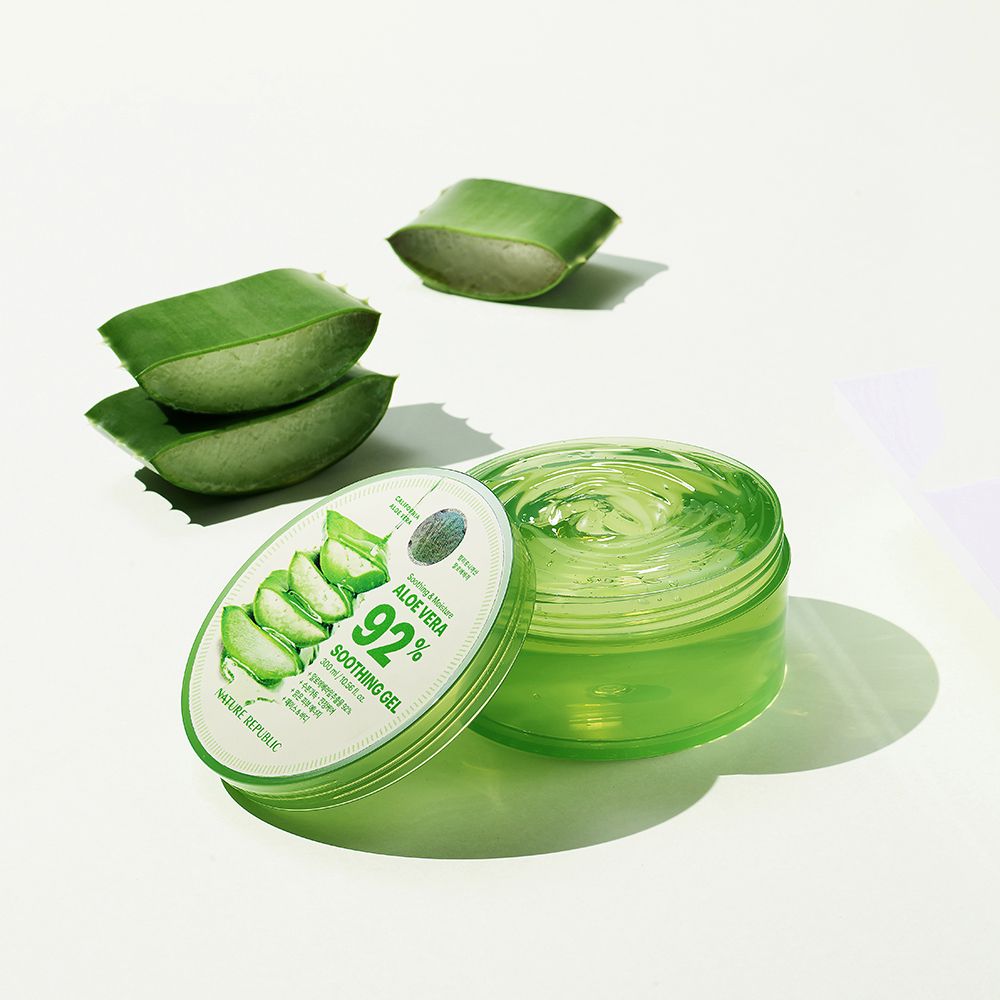 Groene gel in een transparante pot met deksel. Naast, aloë vera bladeren. Op de deksel staat "Aloe Vera 92% Soothing Gel".