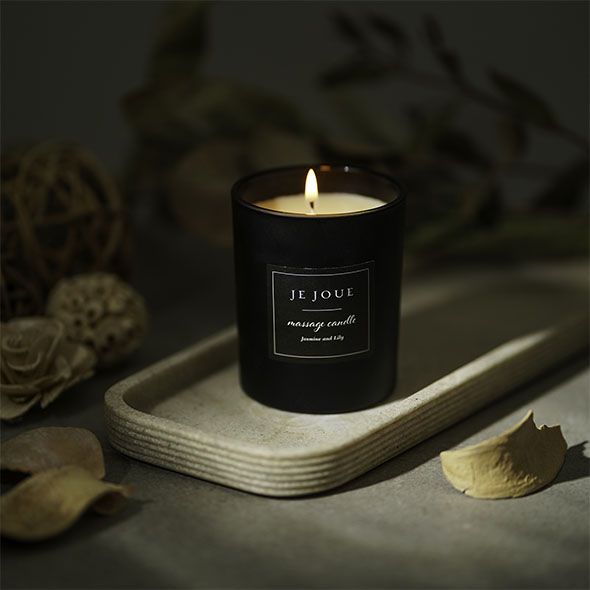 Bougie noire allumée sur un plateau. Inscription : Je Joue, massage candle, Jasmine Lily. Décoration en arrière-plan.