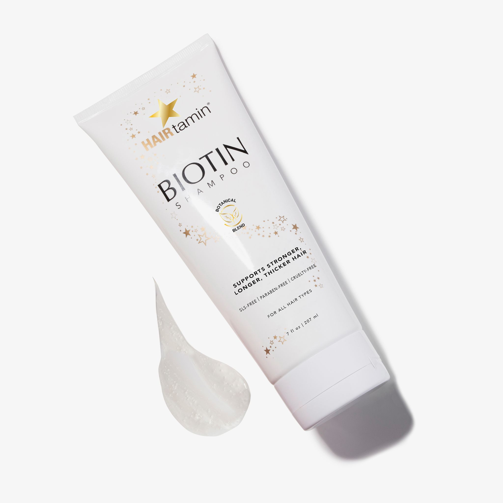 Tube blanc "HAIRtamin Biotin Shampoo" avec coulée. Étoiles et texte "Botanical Blend". Texte : "Supports stronger, longer, thicker hair".