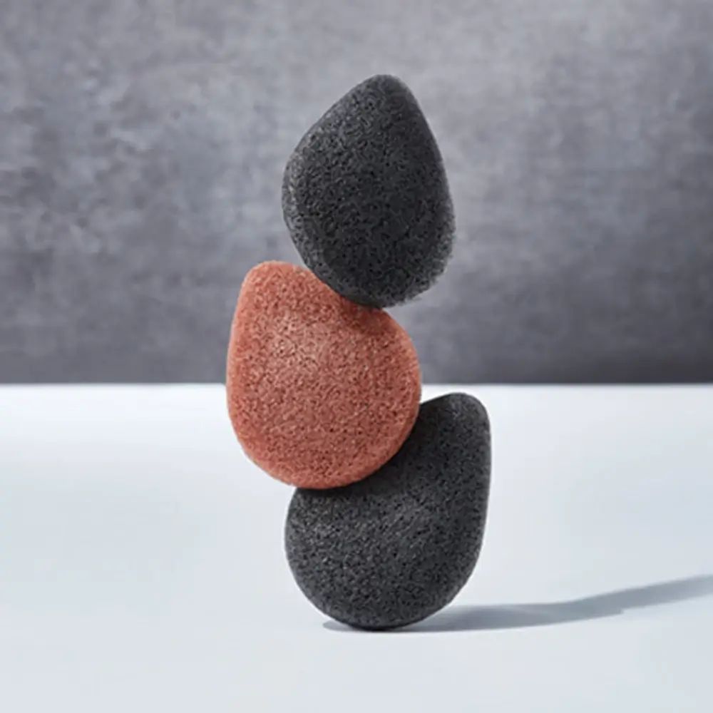 Trois éponges Konjac, deux grises, une brun-rouge, empilées en équilibre. Fond: blanc et gris.