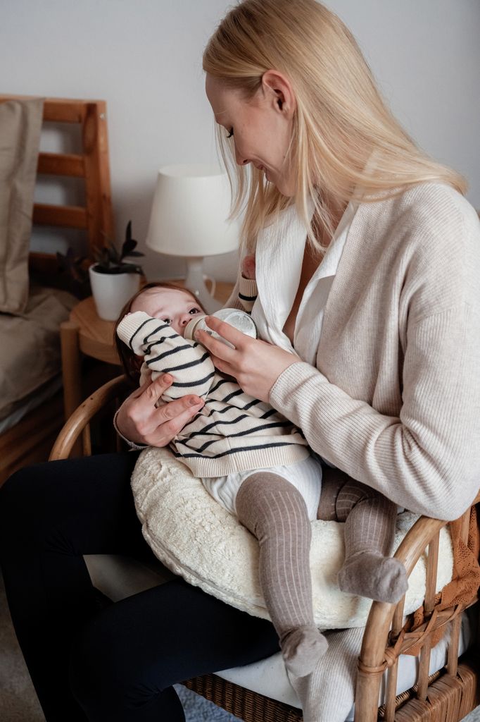 Femme nourrit bébé au biberon, assise sur une chaise avec coussin crème. Bébé porte des vêtements rayés.