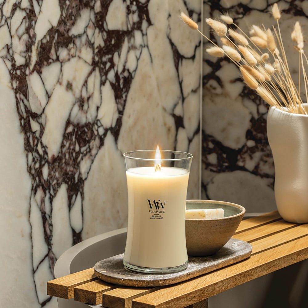 Bougie allumée. Logo WoodWick. Sur un plateau en bois, à côté d'un bol. Fond: marbre.