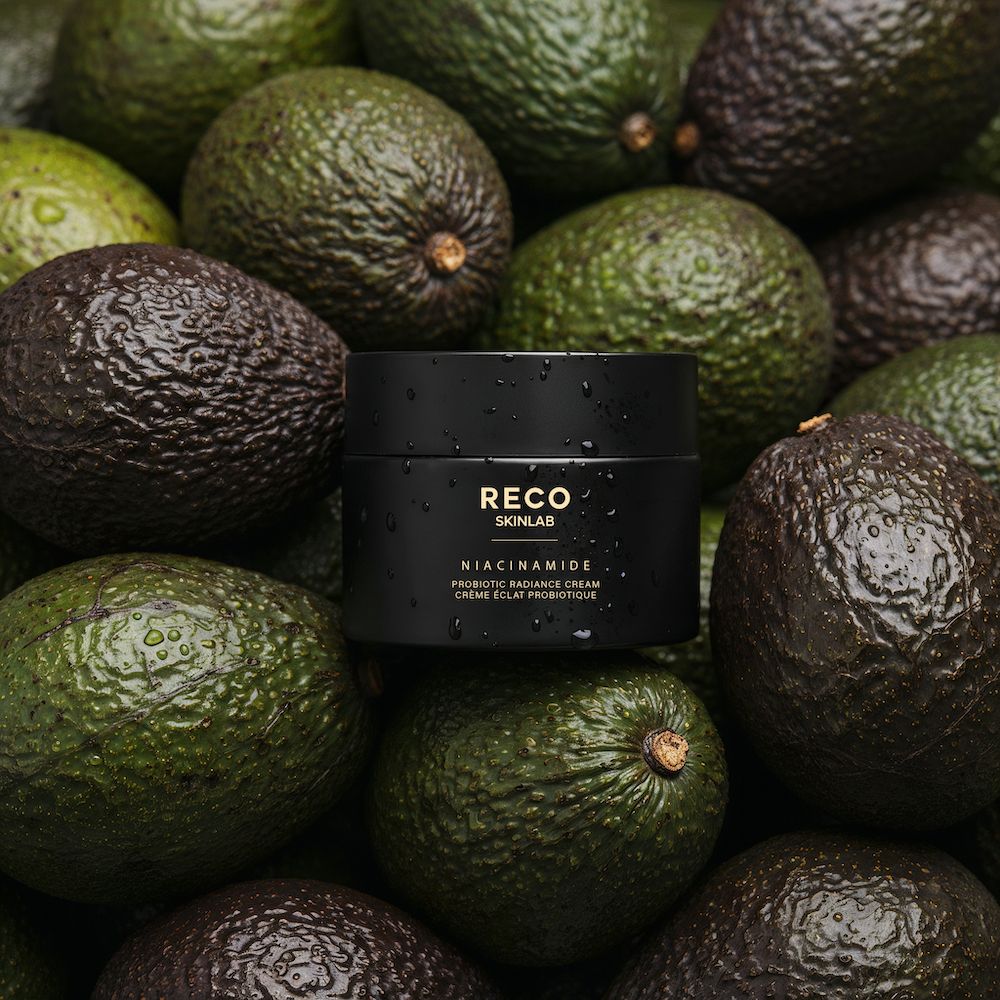 Zwarte pot op avocado's. Opschrift: RECO SKINLAB, Niacinamide Probiotic Radiance Cream. Groene achtergrond.