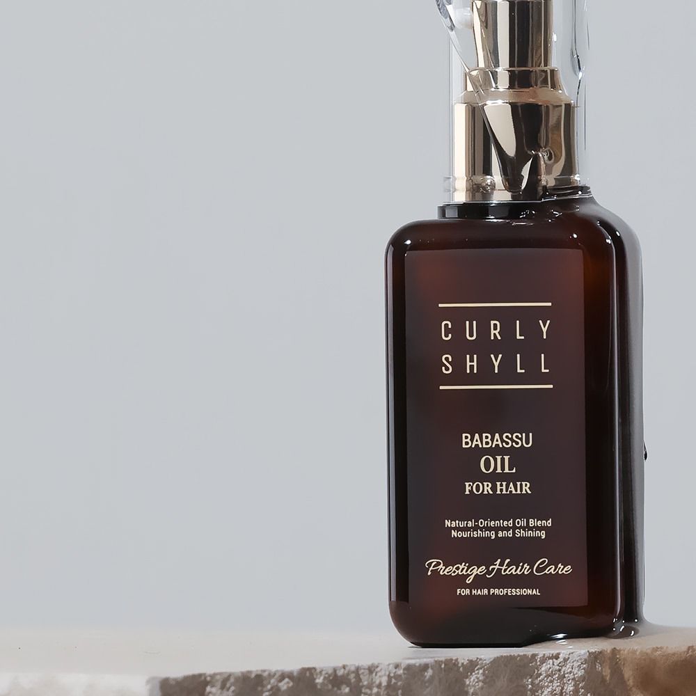 Bruine fles met gouden verstuiver. Opschrift: CURLY SHYLL, Babassu Oil for Hair. Prestige Hair Care.