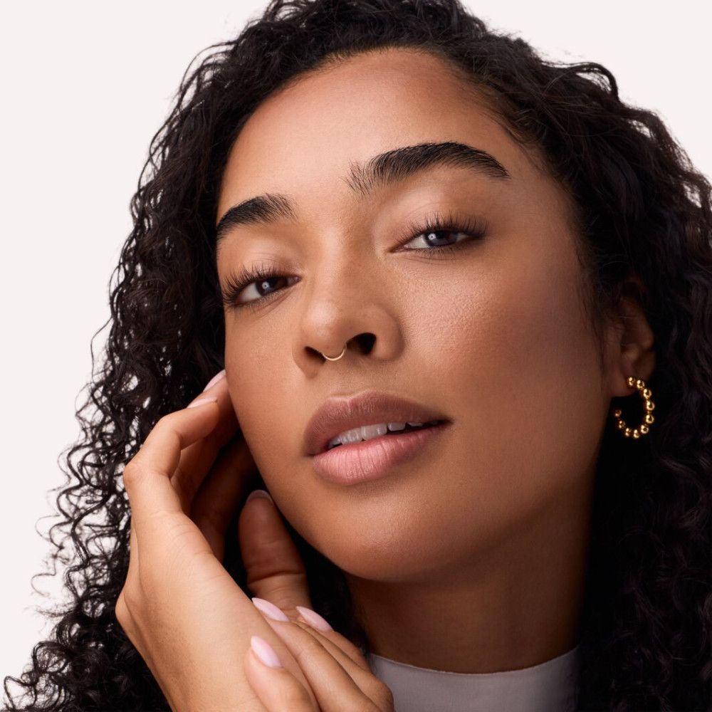 Close-up van een gezicht. Gebronsde huid, donkere krullen, neusring, gouden oorbel. Hand raakt wang aan. Natuurlijk licht.