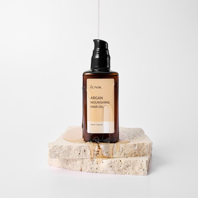 Bruine fles met zwarte pomp op steen. Olie druppelt. Etiket met productnaam en merk. Tekst: Argan Nourishing Hair Oil.