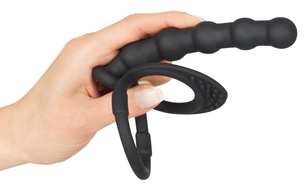 Hand houdt zwarte penis- en ballenring met anale dildo vast. Ring met nopjes, dildo met ribbels.