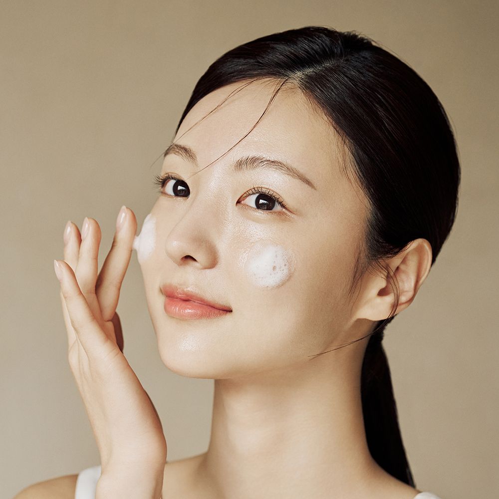 Une femme applique de la mousse sur son visage. Elle touche sa joue avec sa main. La mousse est blanche. En arrière-plan, un fond clair.