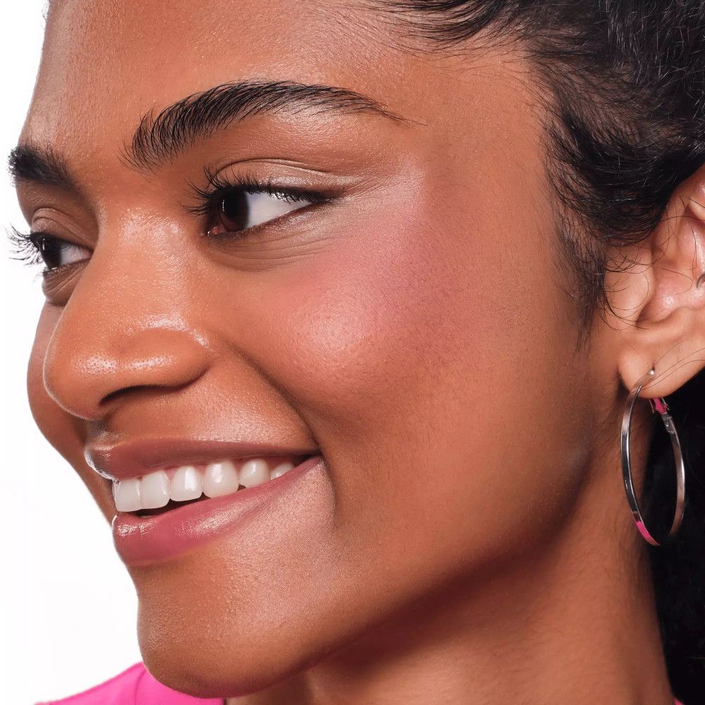 Gros plan d'une femme avec du blush rose sur les joues. Elle porte des boucles d'oreilles argentées et un haut rose.