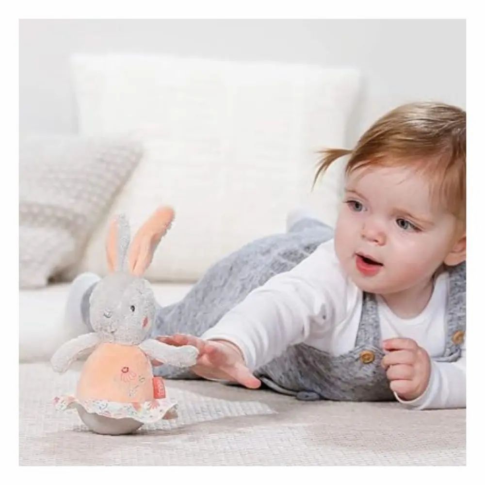 Baby reikt naar een grijs knuffelkonijn met roze buik en oren. Ligt op de grond. Merk: Fehn.