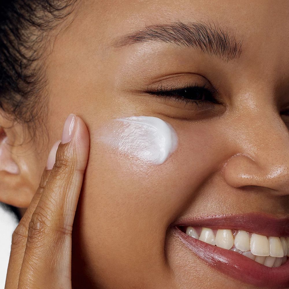 Personne applique une crème blanche sur sa joue. La personne sourit.