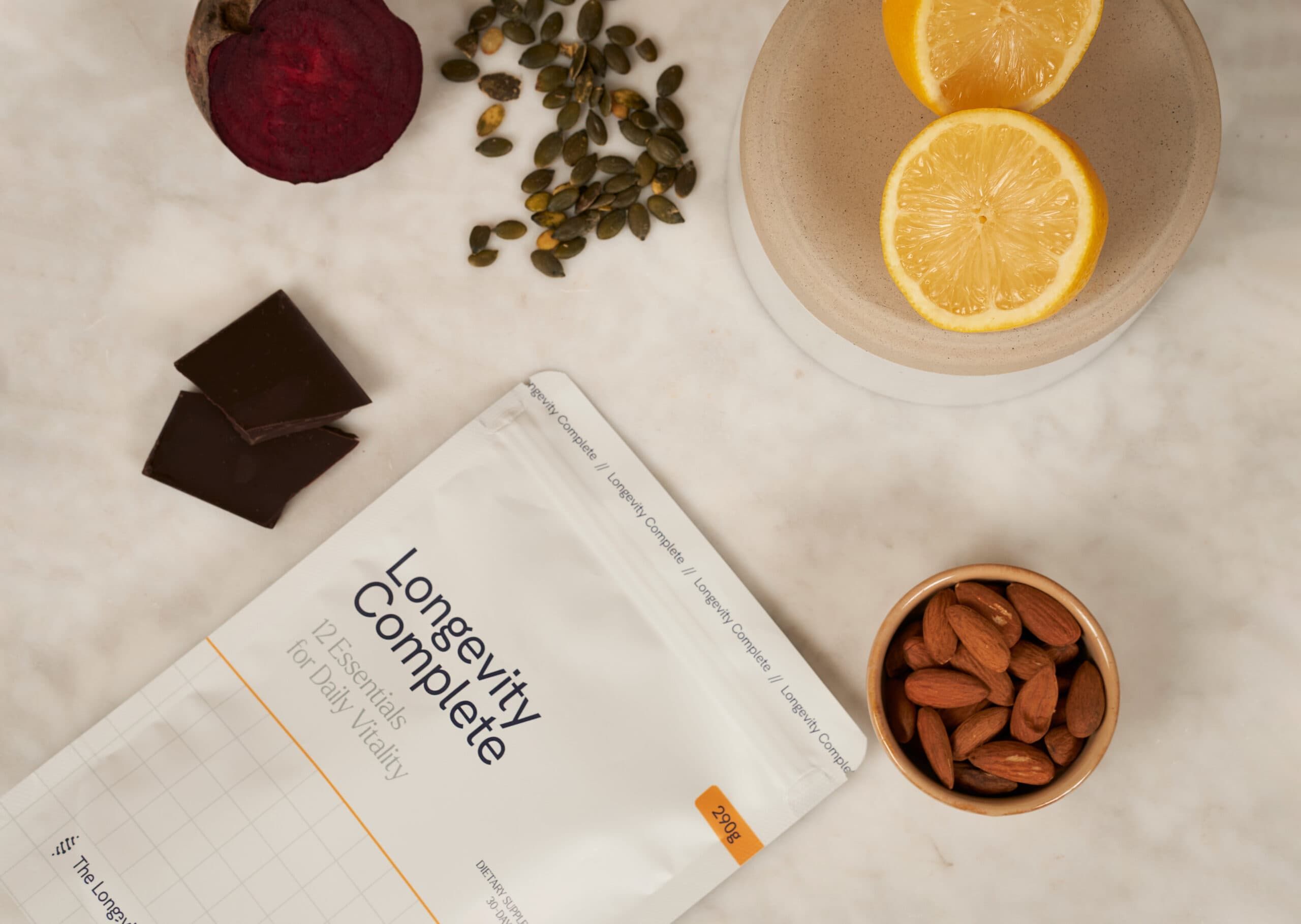 Pochette 'Longevity Complete' avec 12 Essentials pour la vitalité quotidienne. À côté : betterave, graines de courge, chocolat, tranches de citron et amandes.