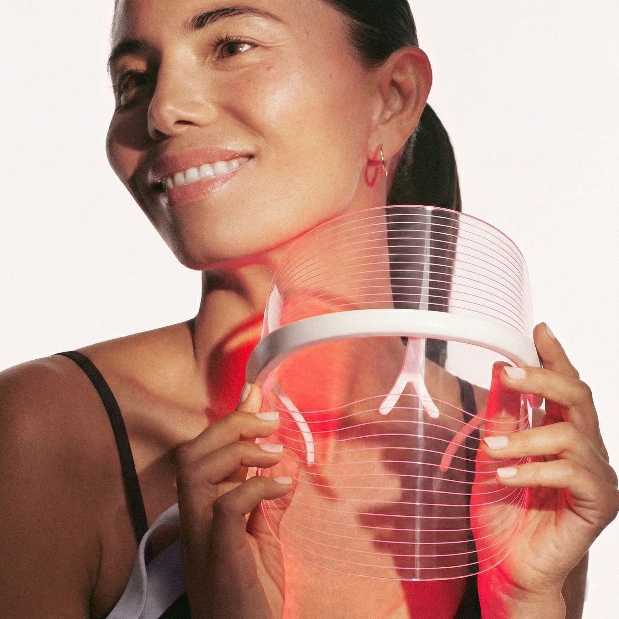 Une femme tient un masque facial LED blanc et transparent, avec des lumières rouges dirigées vers sa zone de cou.