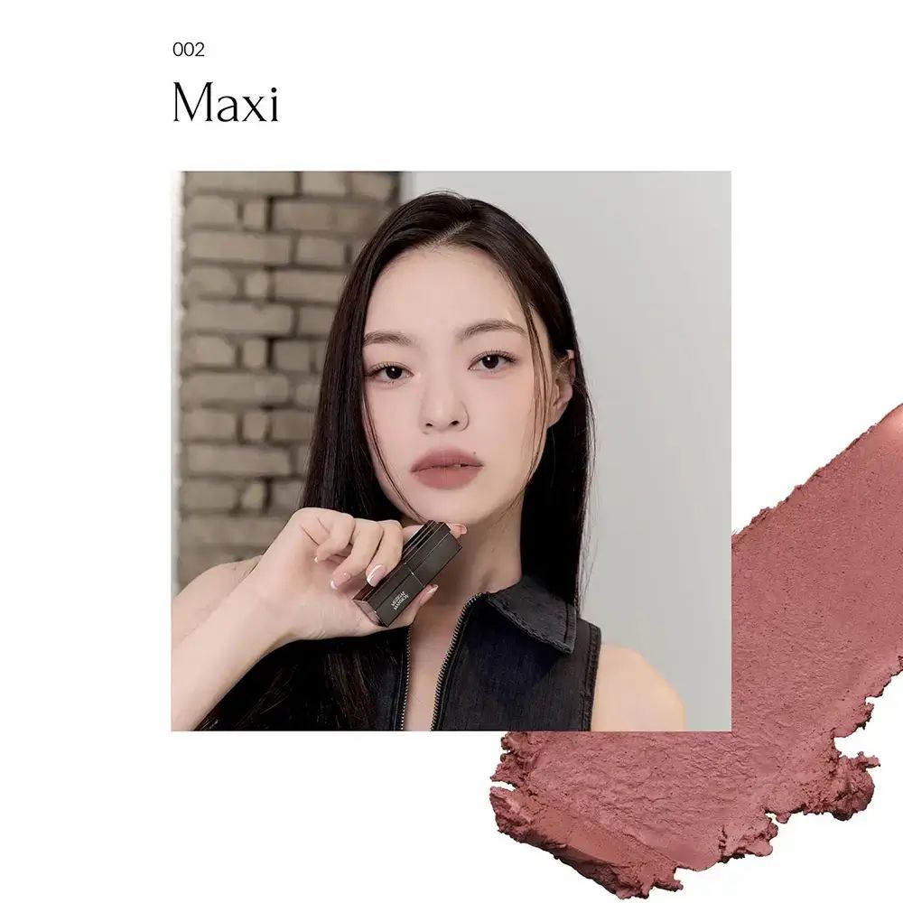 Vrouw houdt lipstick 002 Maxi vast. Lipstick aangebracht op lippen. Kleurveeg ernaast. Achtergrond: bakstenen muur.